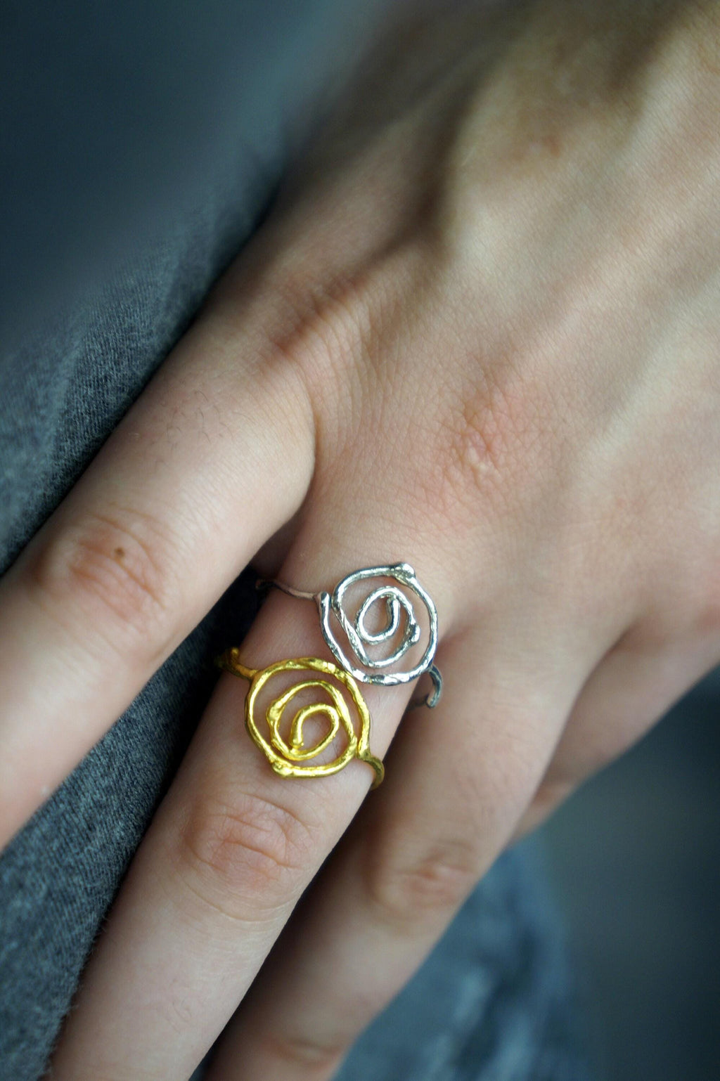 A close-up of a person's hand wearing two rings, one with a gold spiral design and the other with a silver spiral design.