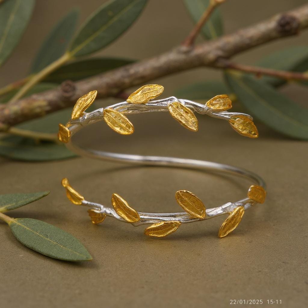 A silver bracelet with gold leaf-shaped charms is displayed on a branch with green leaves, creating a natural and elegant composition.