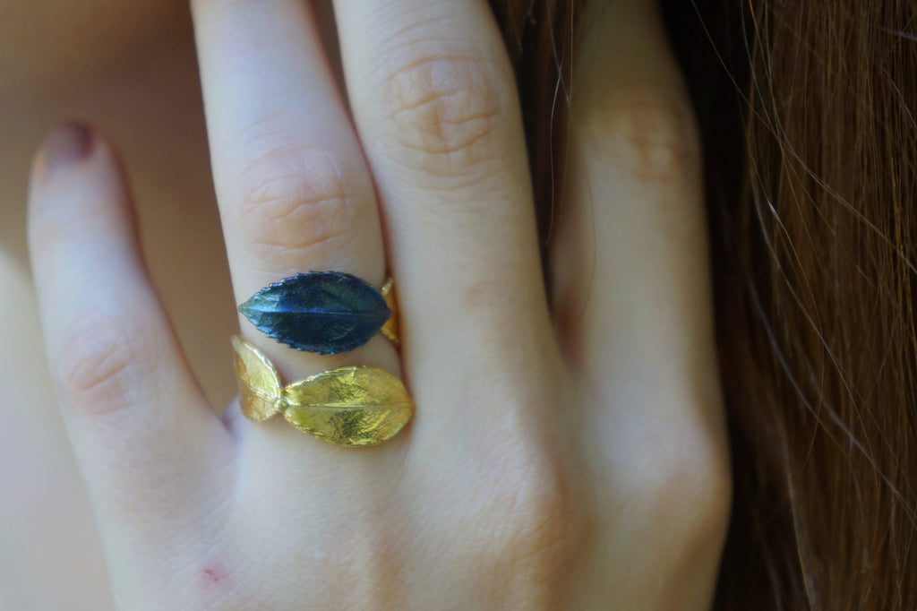A close-up of a person's hand wearing a ring with two leaf-shaped gemstones, one blue and one gold.