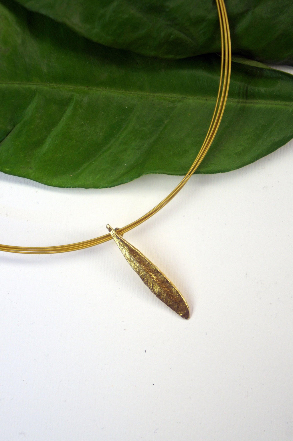 A gold-colored pendant necklace with a leaf-shaped pendant is displayed against a white background, with a green leaf partially visible behind the necklace.