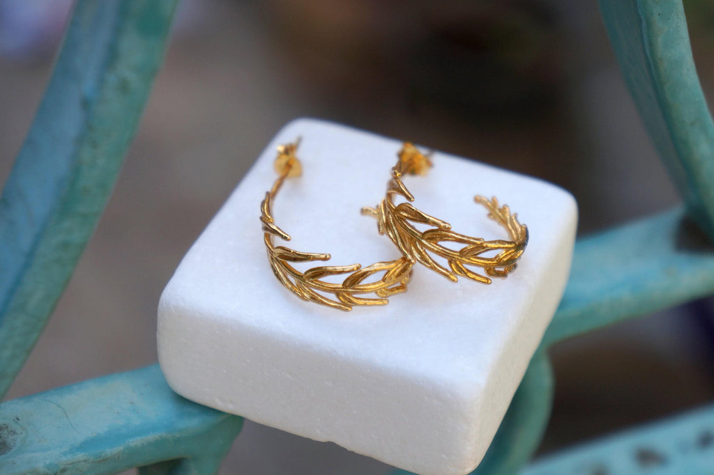 A pair of gold earrings with leaf-like designs rests on a white surface, with a blurred background.
