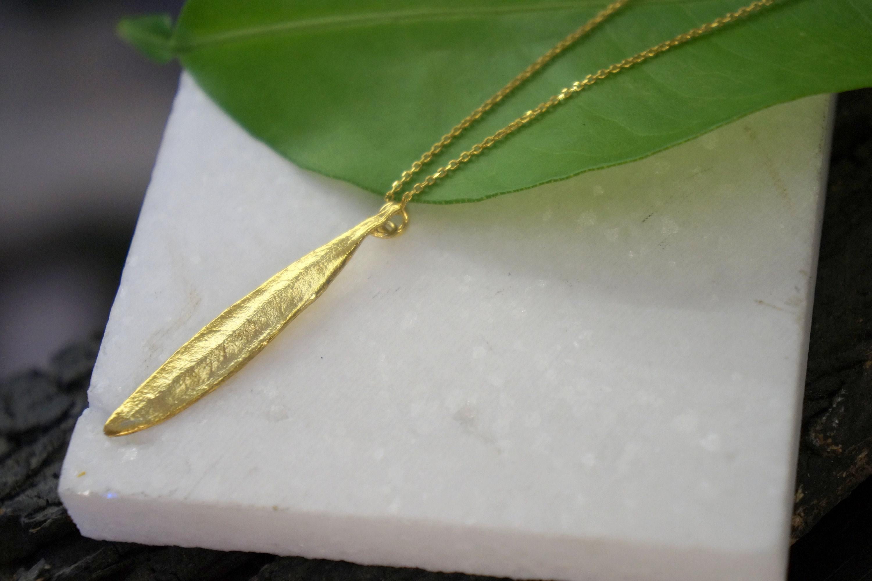A gold necklace with a leaf pendant is displayed on a white marble surface, with a green leaf partially covering the necklace.
