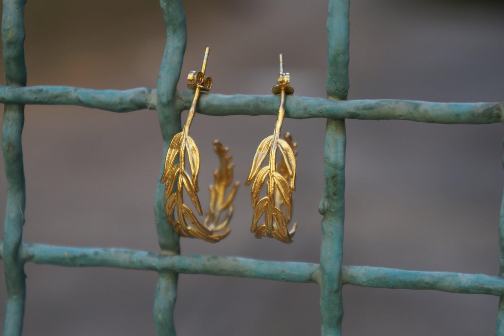 A pair of gold earrings with leaf-shaped designs hanging from a metal fence.