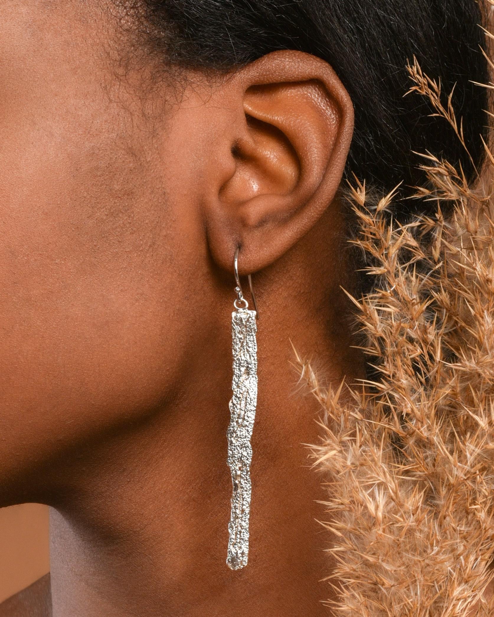 A close-up view of a person's ear wearing a silver earring with a long, thin, metallic design.