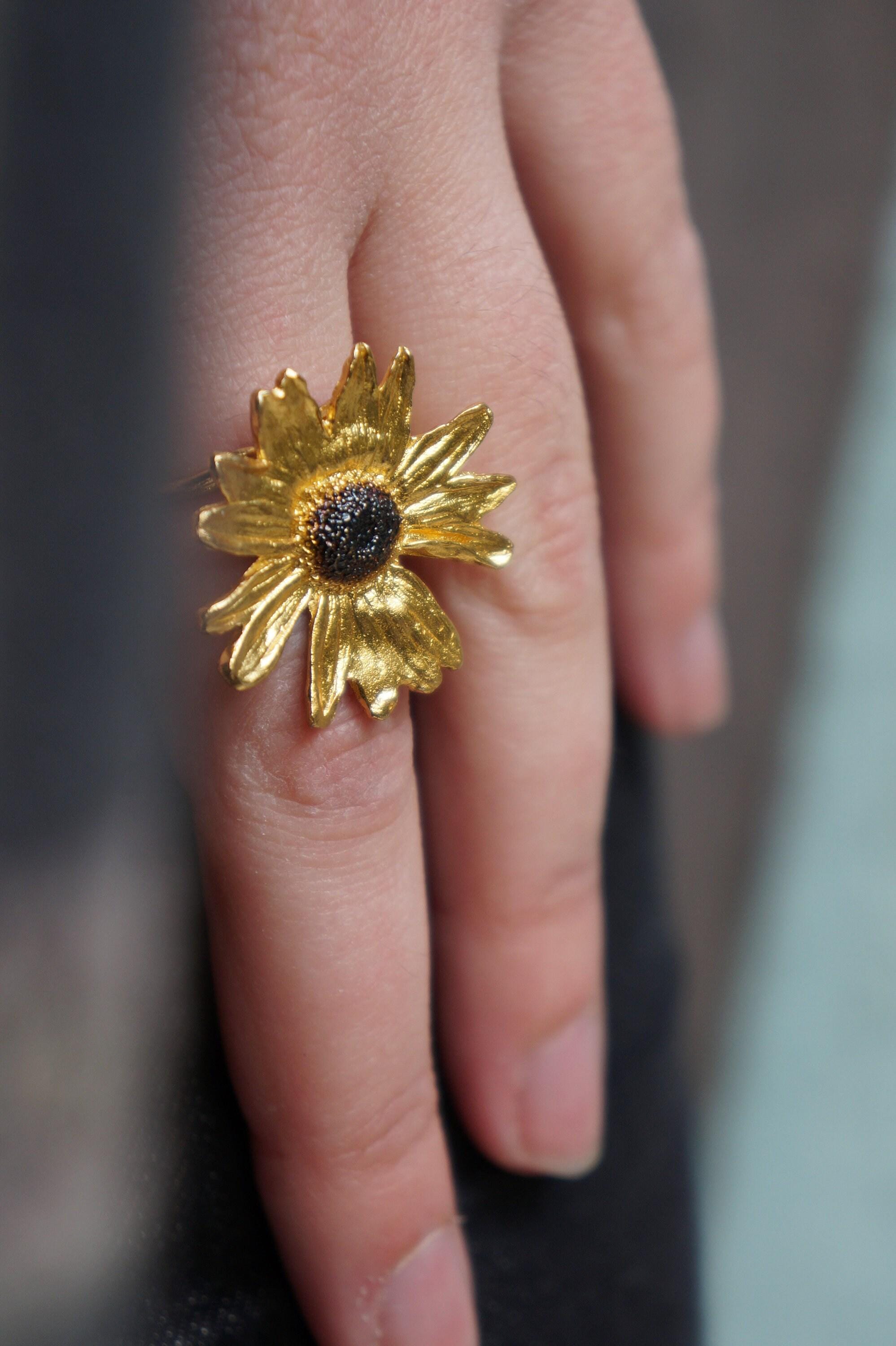 A close-up of a person's hand wearing a gold ring with a large, intricately designed flower as the centerpiece.