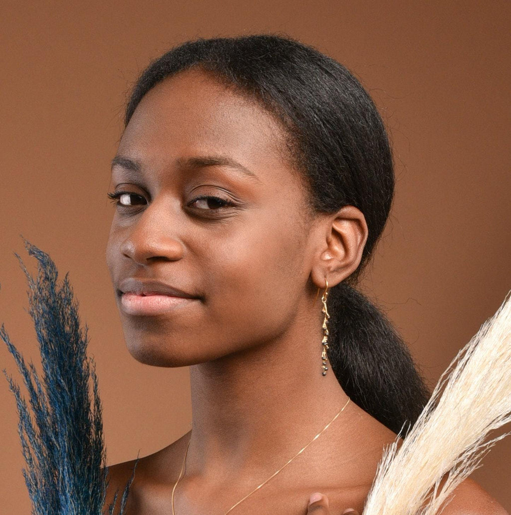 A portrait of a young woman with dark hair wearing gold earrings and a necklace, holding a blue and white flower.