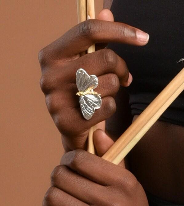 A close-up of a person's hand holding a silver butterfly-shaped ring, with wooden sticks in the background.
