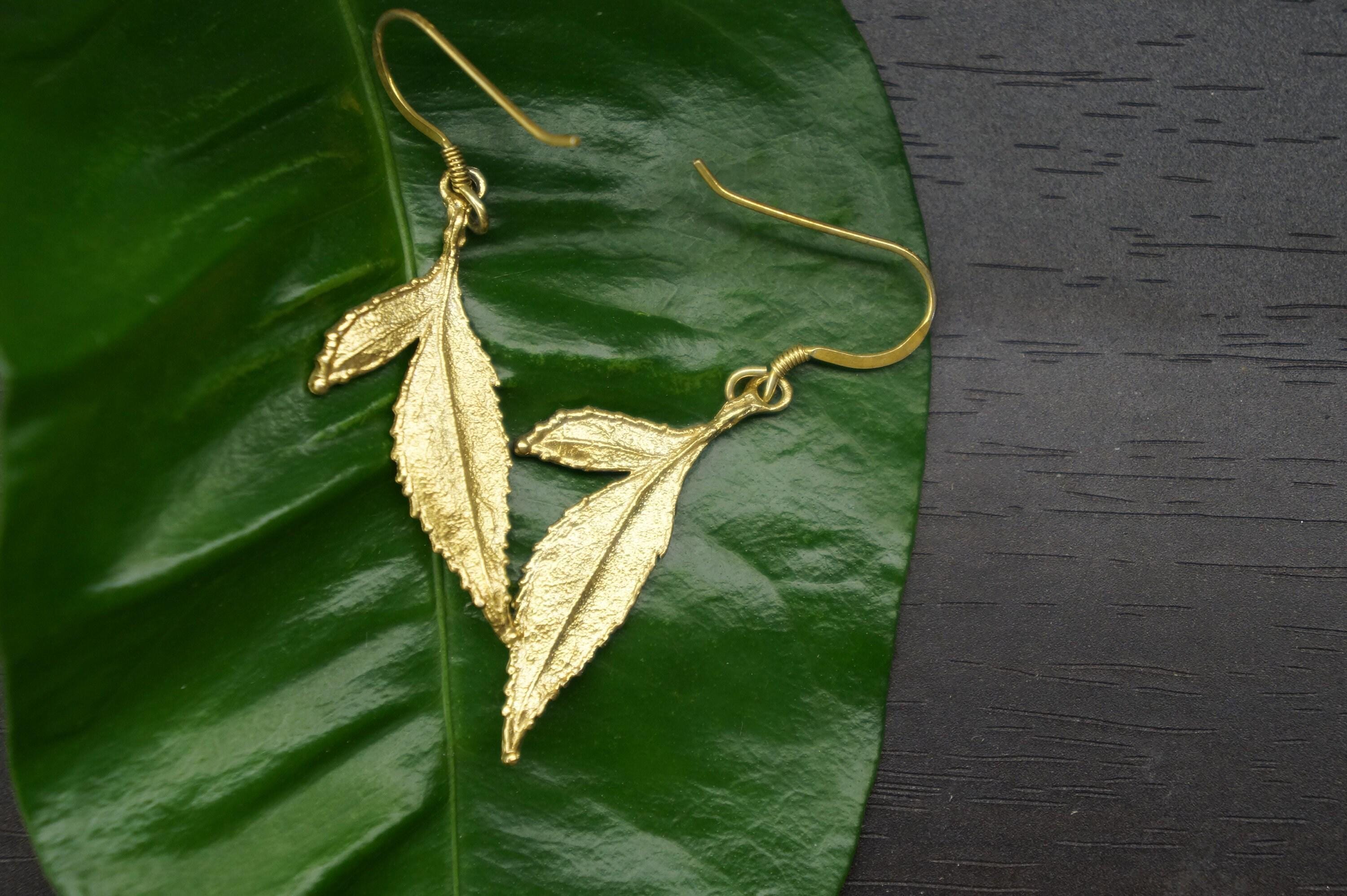 A pair of gold leaf earrings rests on a green leaf against a dark background.