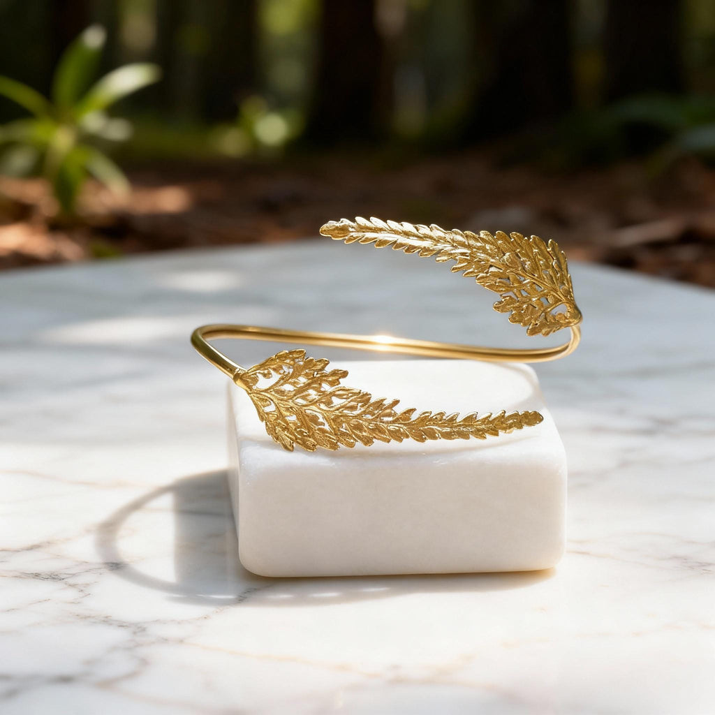 A gold bracelet with leaf-shaped designs rests on a white marble surface, with a blurred natural background.