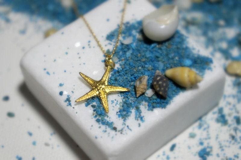 A gold starfish pendant is displayed on a white surface, surrounded by various seashells and sand.
