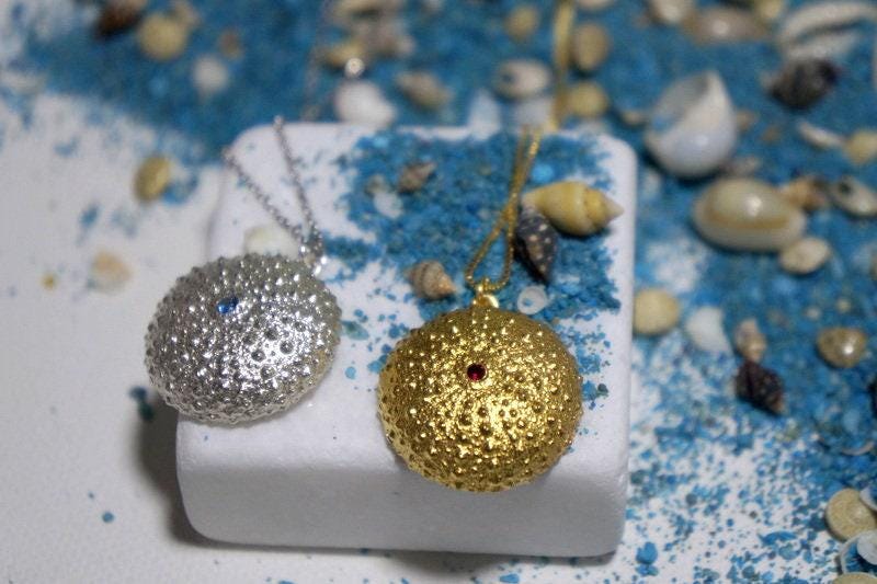 two round pendants, one silver and one gold, resting on a white surface with scattered seashells and sand.