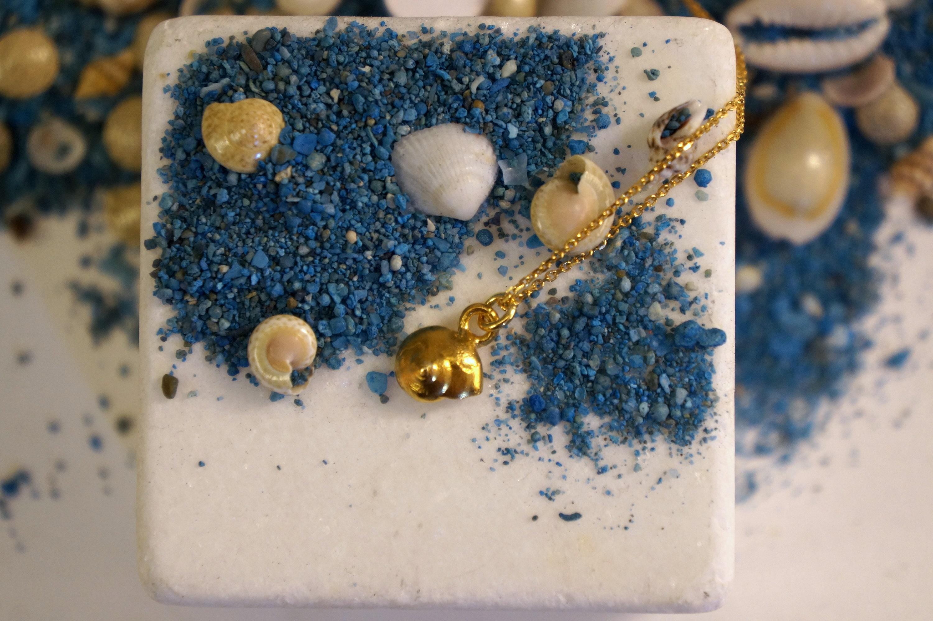 A white square-shaped object with a gold-colored charm hanging from it, surrounded by various seashells and sand in shades of blue and white.