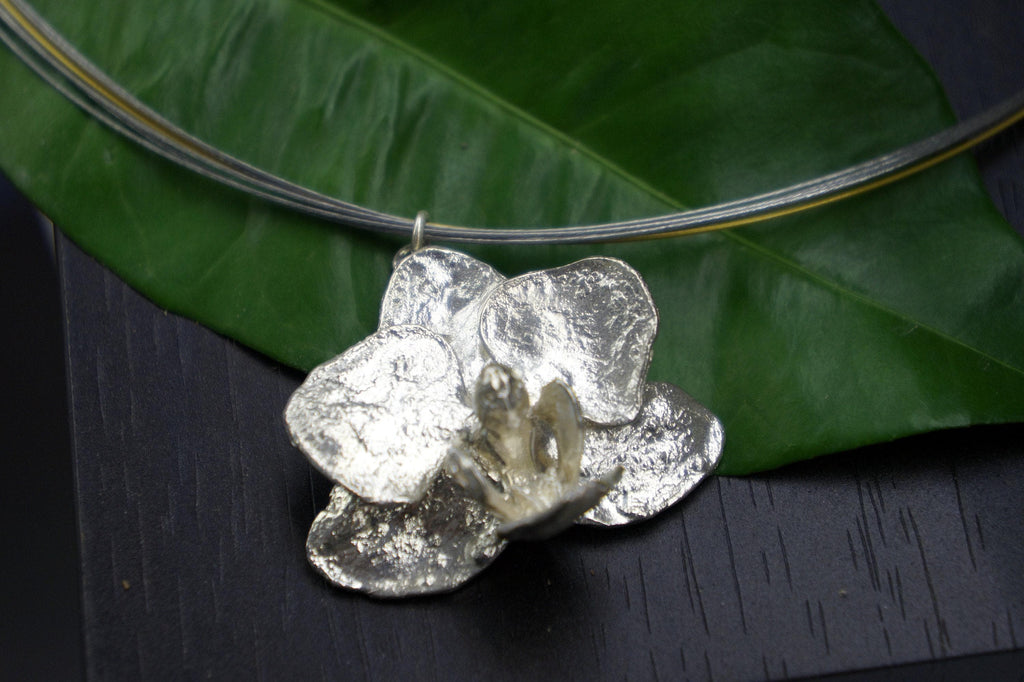 A silver pendant in the shape of a flower is displayed on a green leaf, with a necklace chain visible in the background.