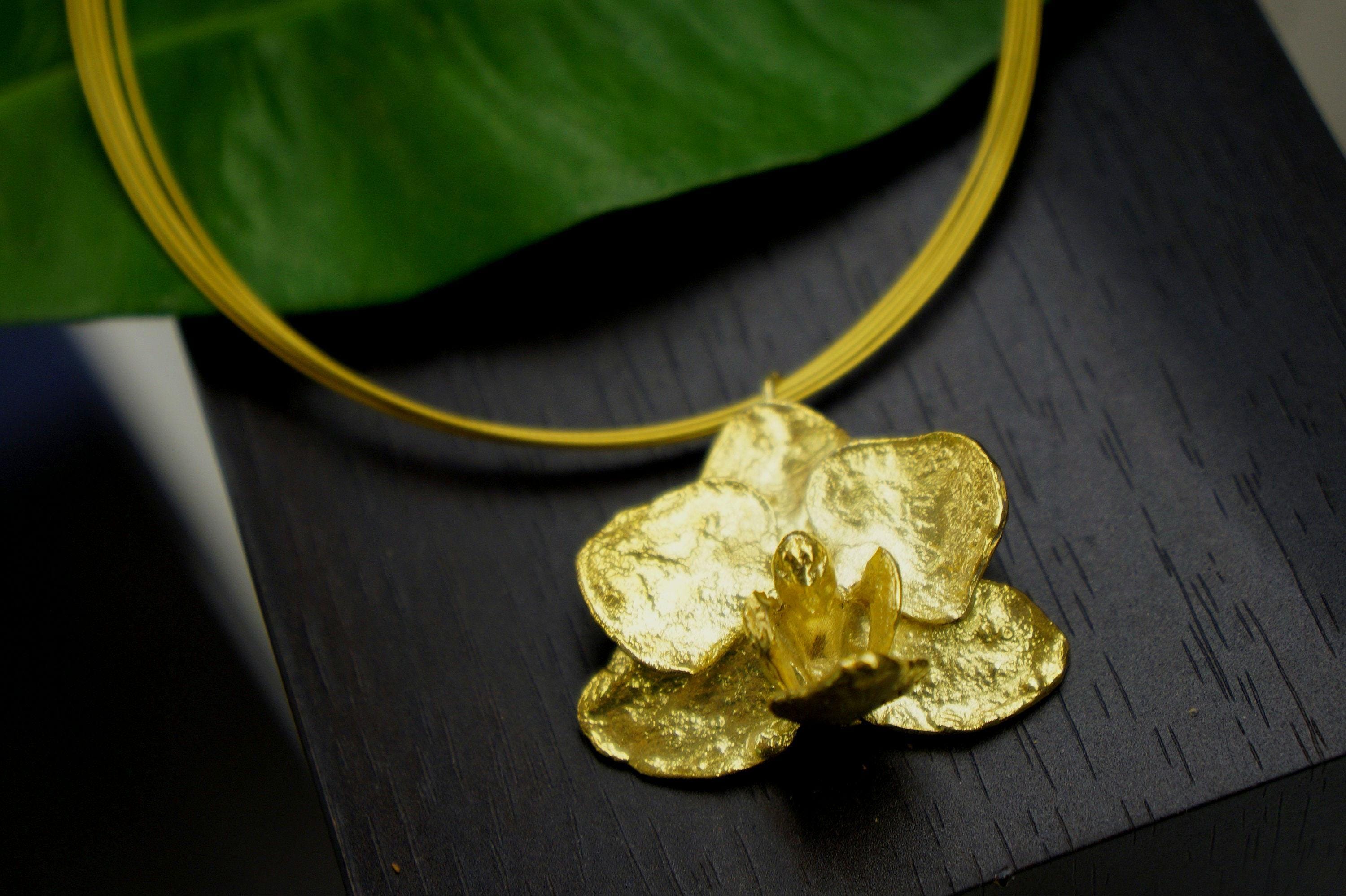 A gold-colored pendant in the shape of a flower is displayed on a black surface, with a green leaf visible in the background.
