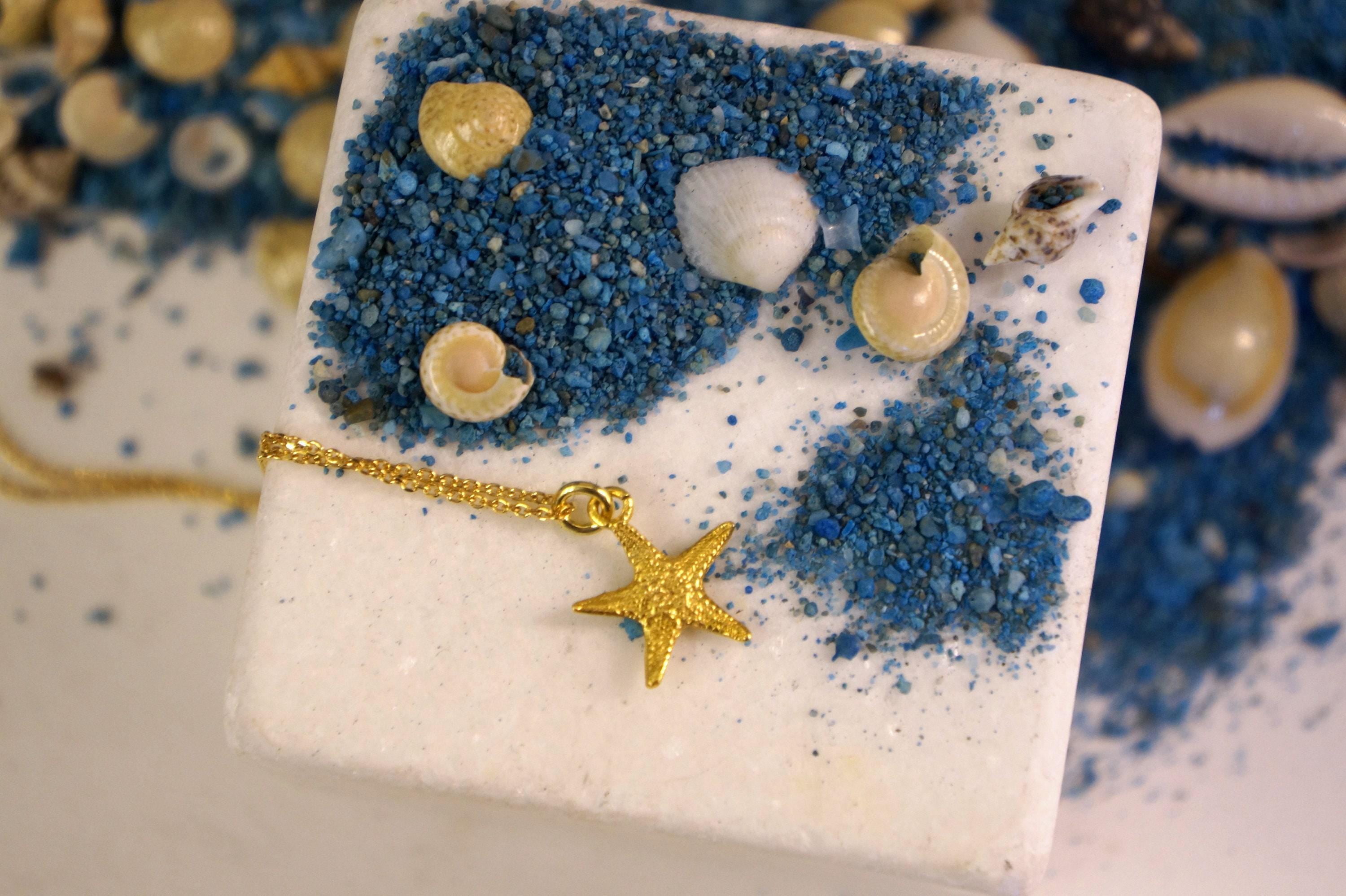 A white square-shaped object with a gold star charm and various seashells and blue sand on top, creating a beach-themed design.