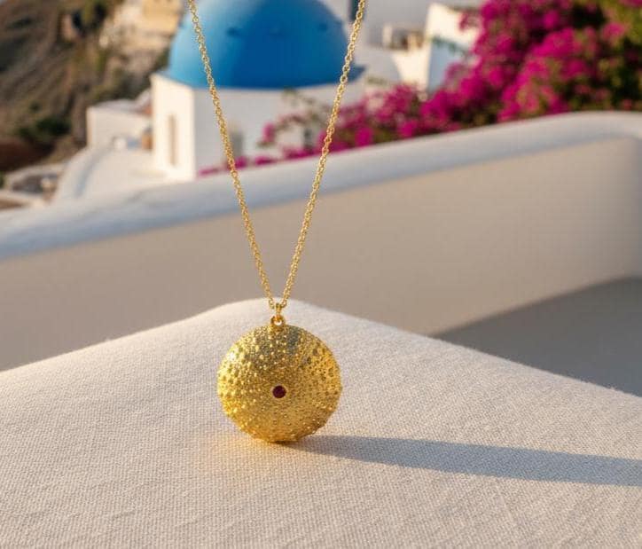 A gold necklace with a round pendant is displayed on a white surface, with a blue dome and pink flowers in the background.