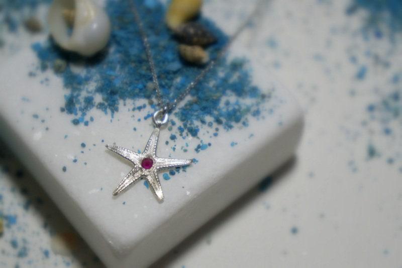 A silver starfish pendant with a red gemstone is displayed on a white surface, surrounded by blue sand and small seashells.