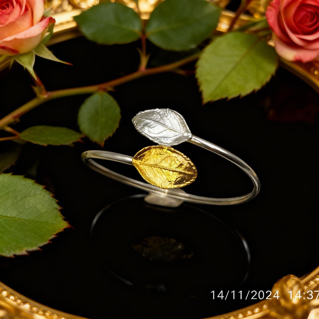 A silver and gold leaf-shaped bracelet with two leaf-shaped charms is displayed on a black background, surrounded by green leaves and pink roses.