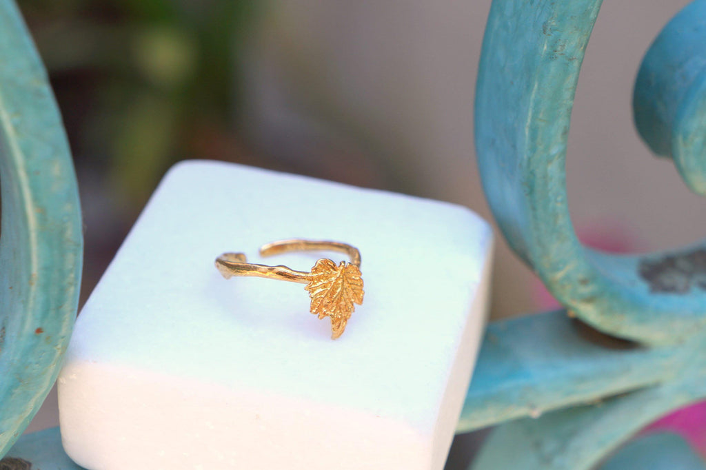 A gold leaf ring is displayed on a white square, placed on a blue metal railing.