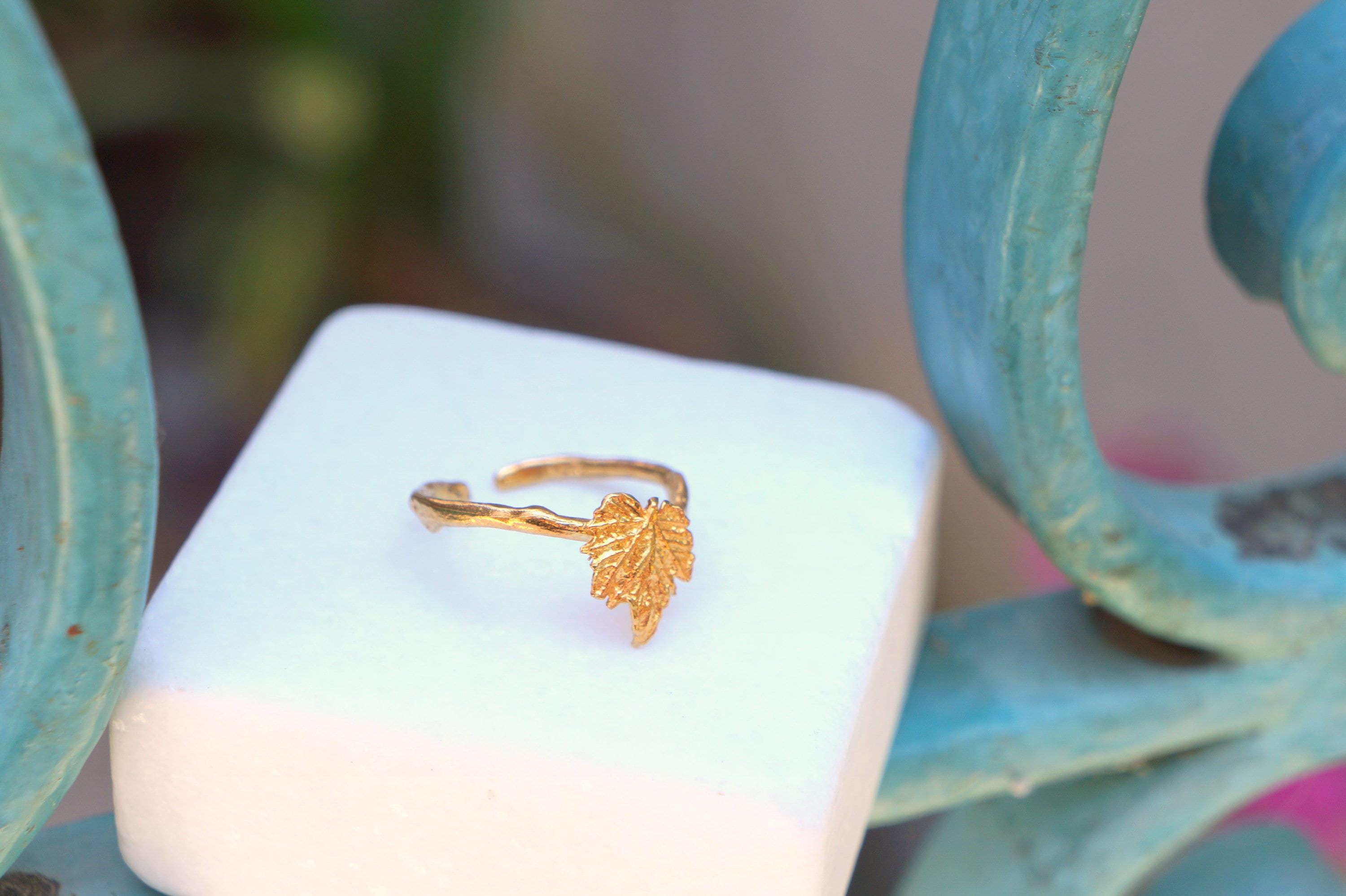 A gold leaf ring is displayed on a white square, placed on a blue metal railing.