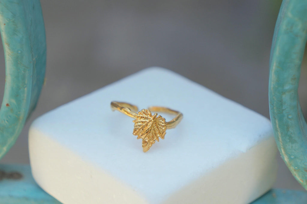 A gold leaf ring is displayed on a white square surface, with a blurred background.