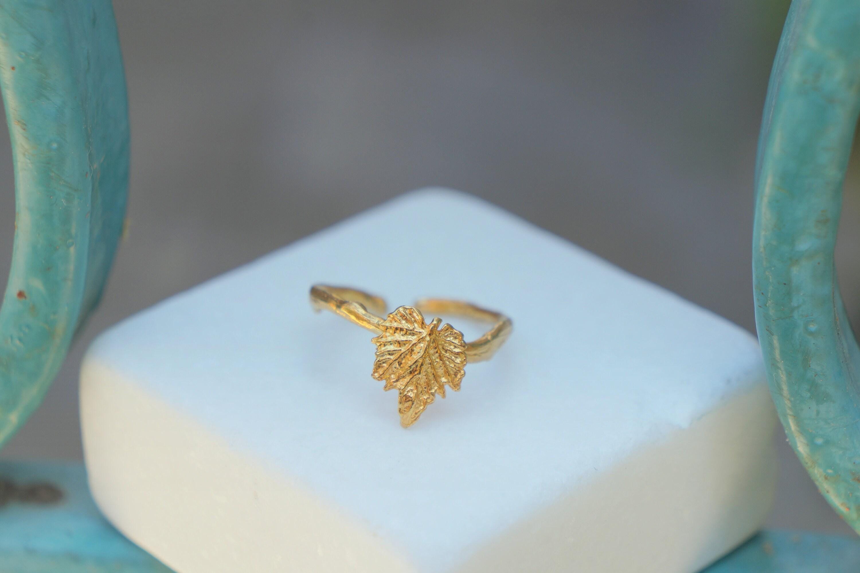 A gold leaf ring is displayed on a white square surface, with a blurred background.