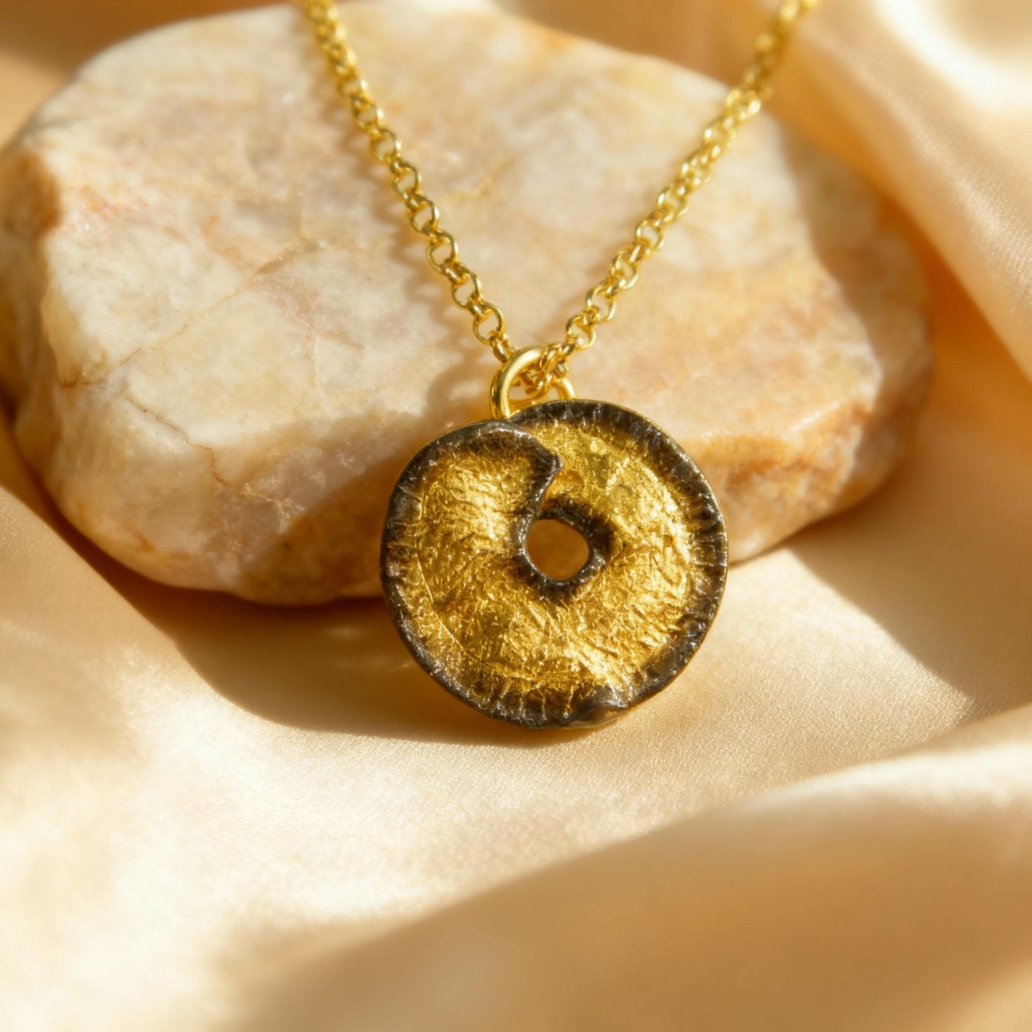 A gold necklace with a circular pendant is displayed on a beige fabric surface.
