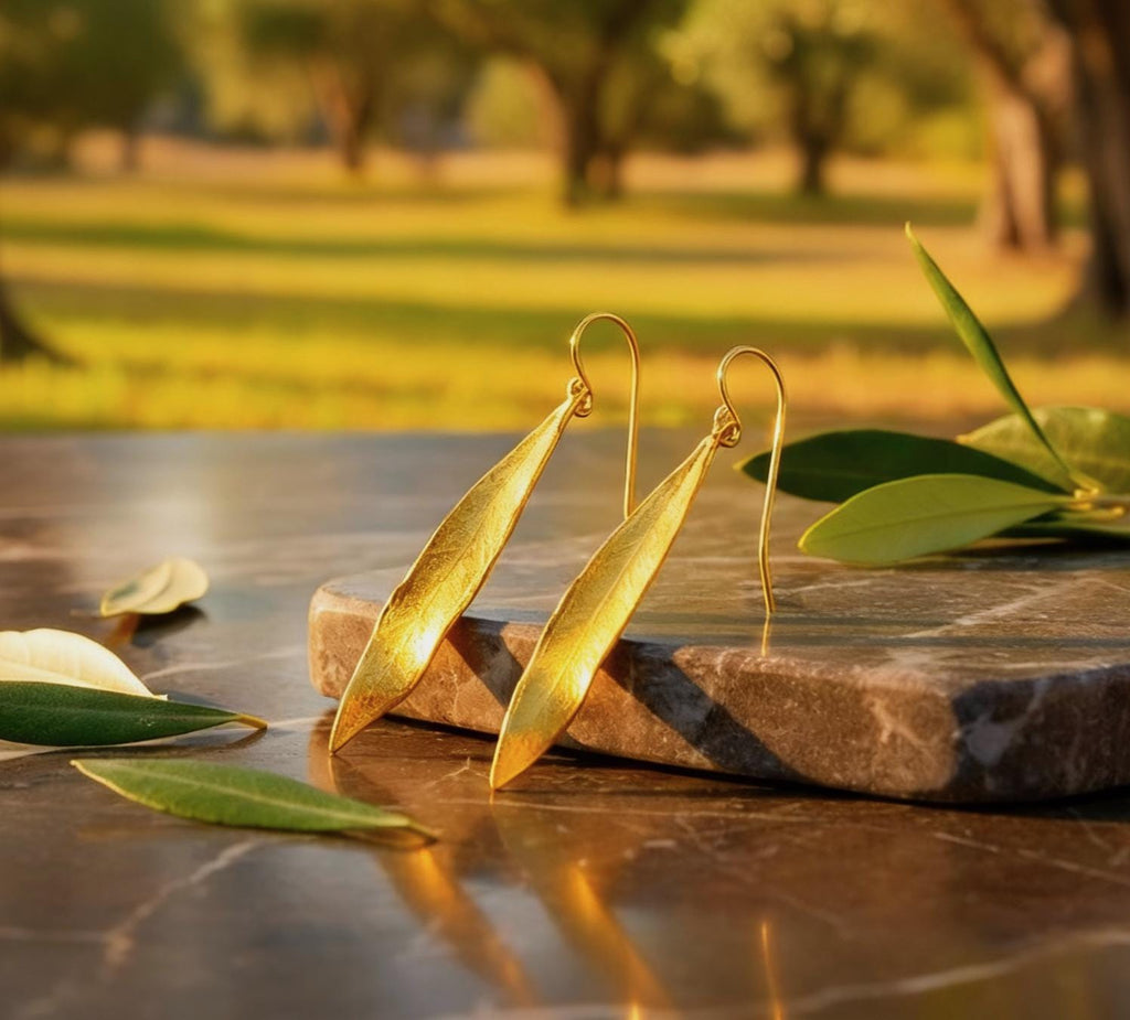 A pair of gold leaf earrings rests on a marble surface, surrounded by fallen leaves, with a blurred background of trees and greenery.