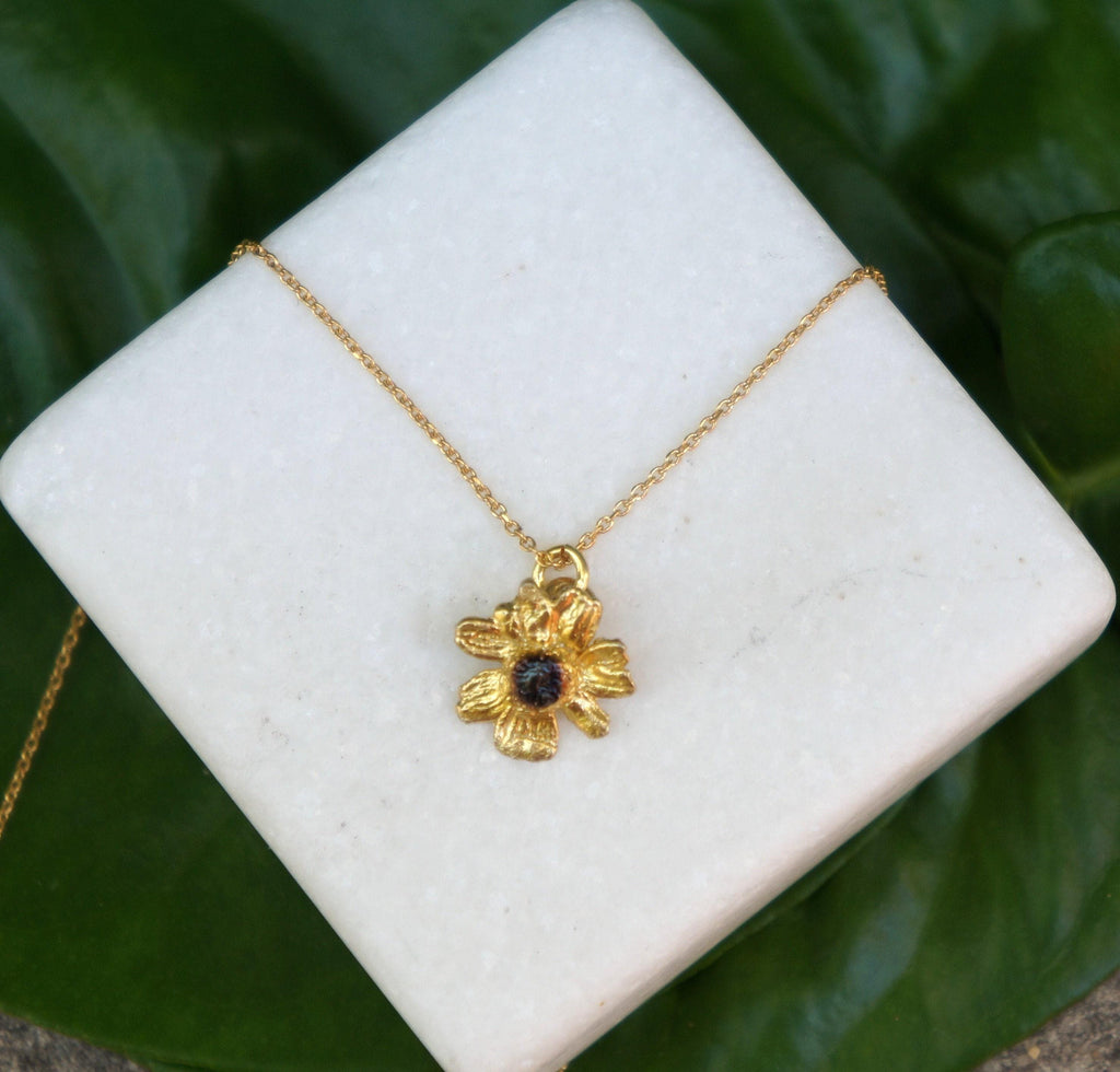 A gold-colored necklace with a pendant featuring a flower design is displayed on a white square surface against a green leaf background.