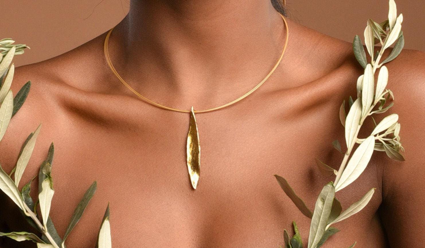 A close-up of a woman's neck and chest area, with a gold necklace featuring a pendant and leaves surrounding her.