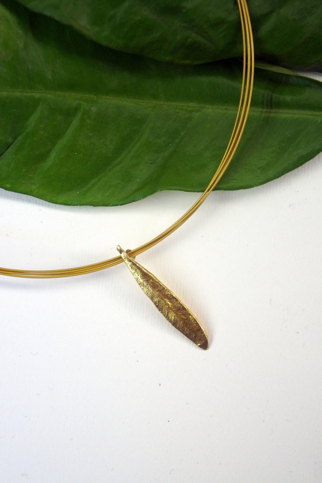 A gold-colored pendant with a leaf-like design is displayed on a white surface, with a gold wire necklace around it.