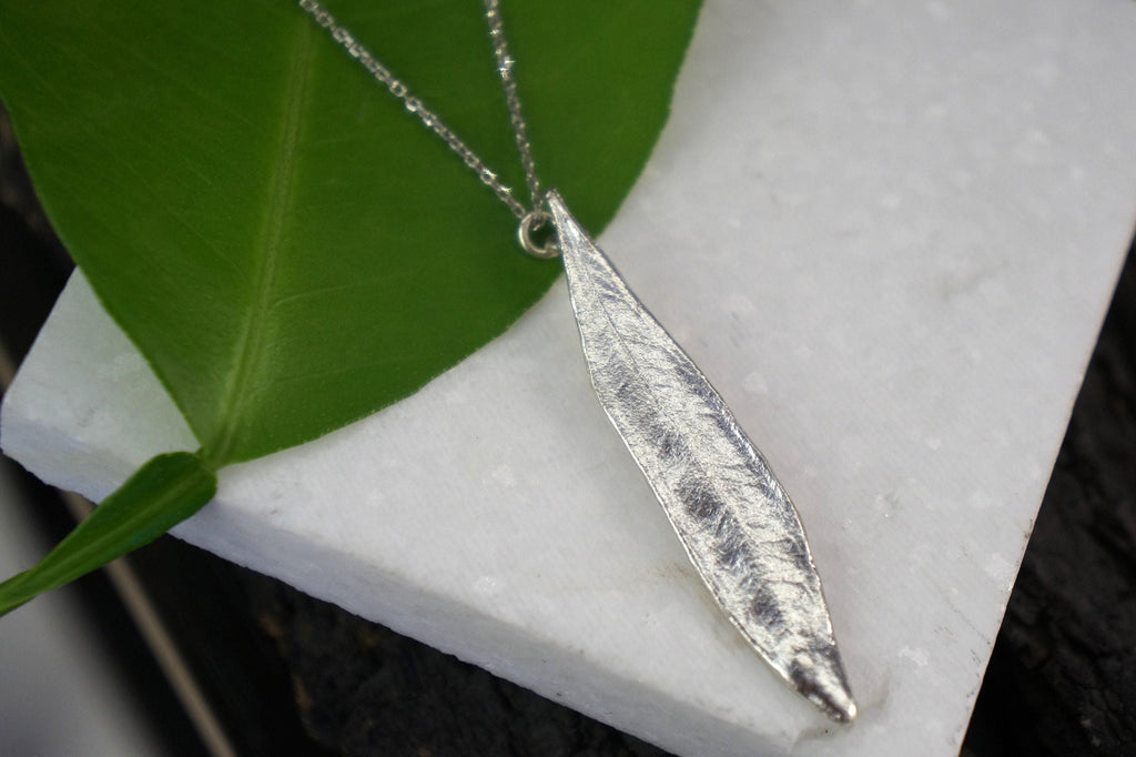 A silver leaf-shaped pendant necklace is displayed on a white marble surface, with a green leaf partially visible to the left.