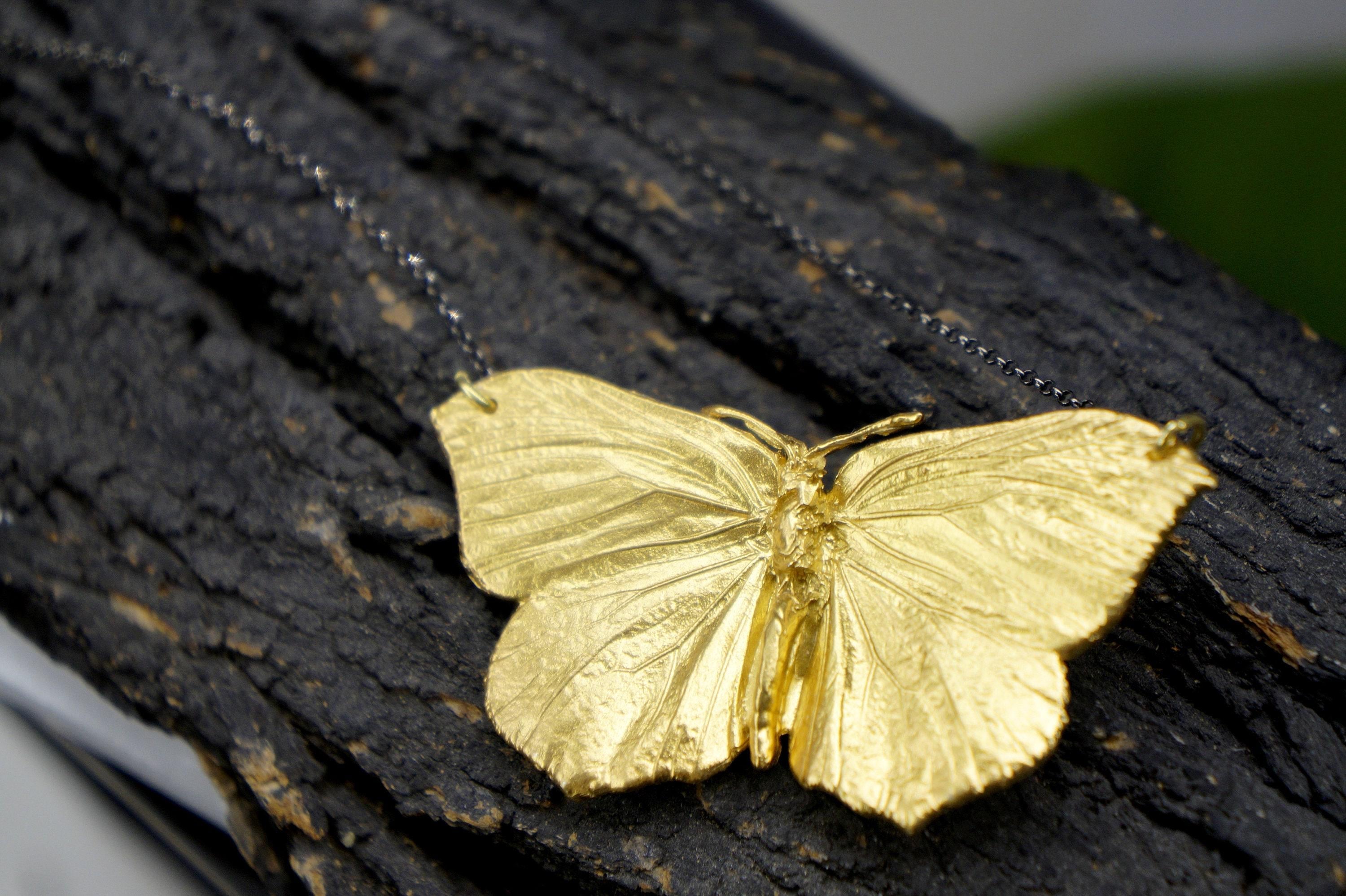 A close-up image of a gold butterfly pendant hanging from a chain against a dark, textured background.