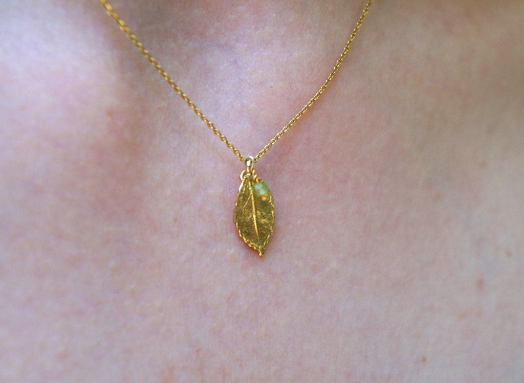 A gold-colored necklace with a leaf pendant is shown against a pinkish-brown background.
