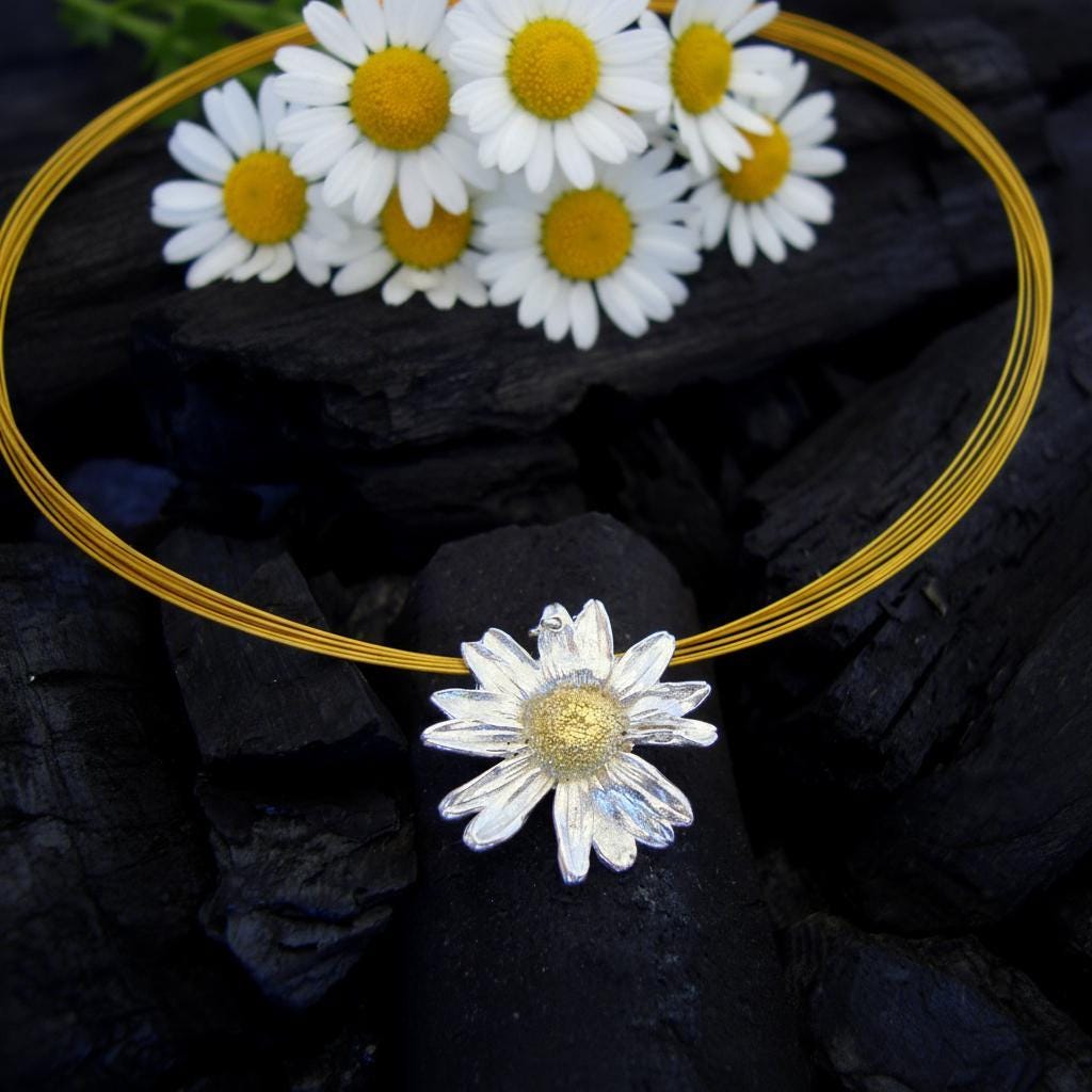 a necklace with a large white daisy pendant, surrounded by a circular arrangement of white and yellow daisies. The daisies are placed on a black surface, creating a striking contrast.