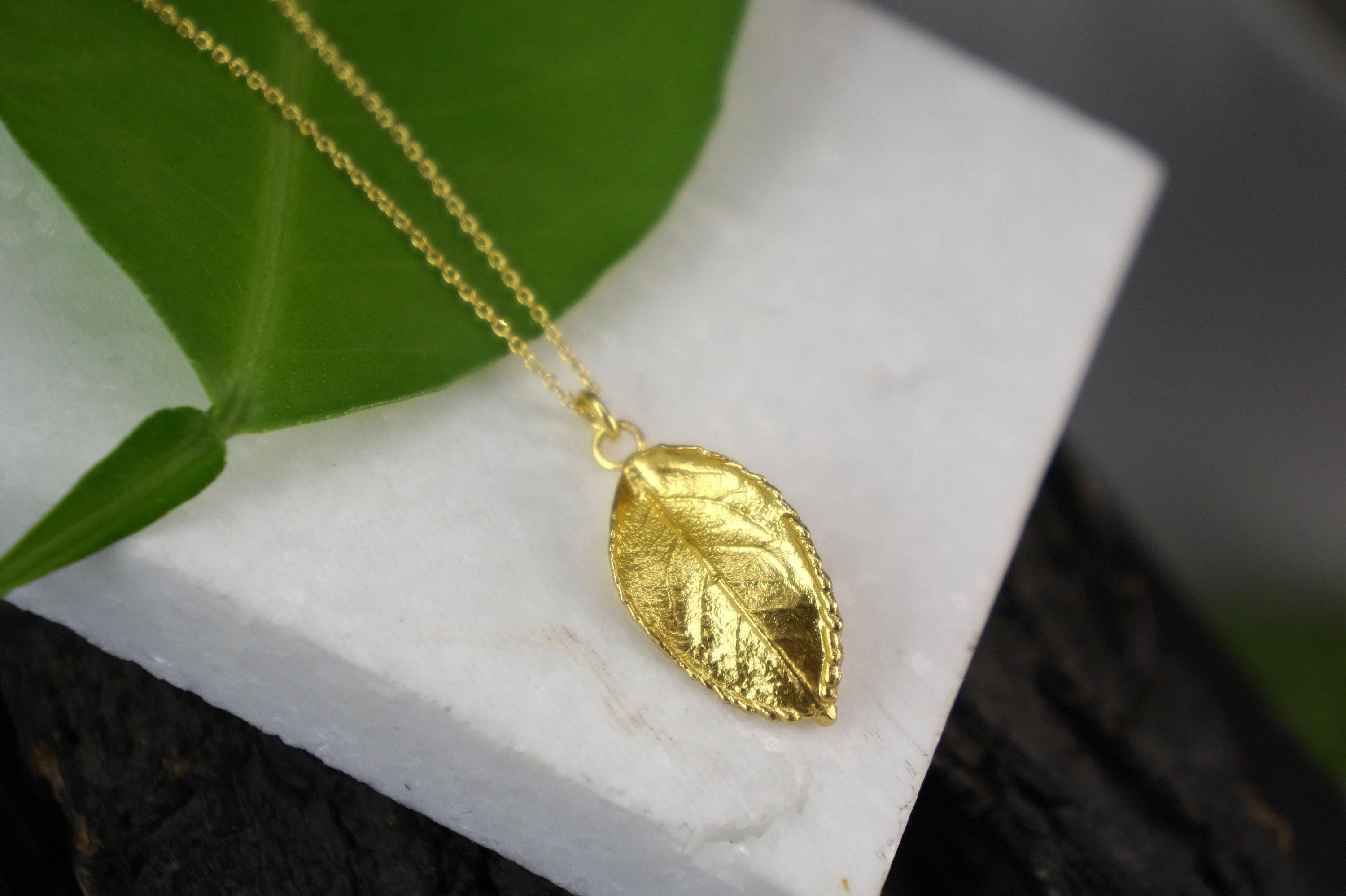 A gold leaf pendant necklace rests on a white marble surface, with a green leaf partially visible behind it.