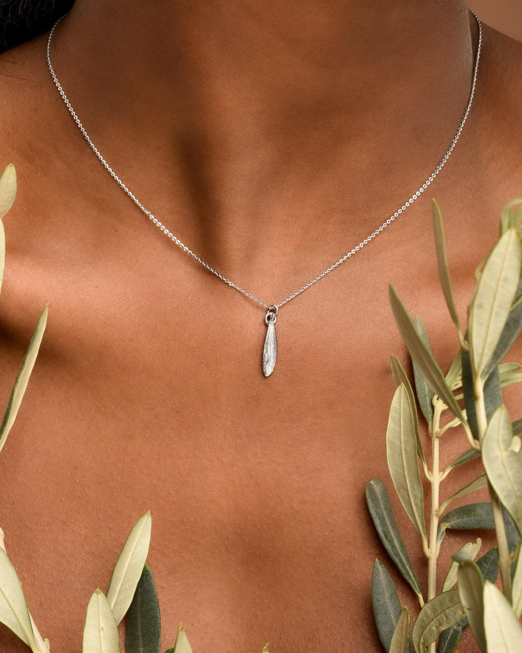 A delicate silver necklace with a small pendant is displayed against a backdrop of green leaves.